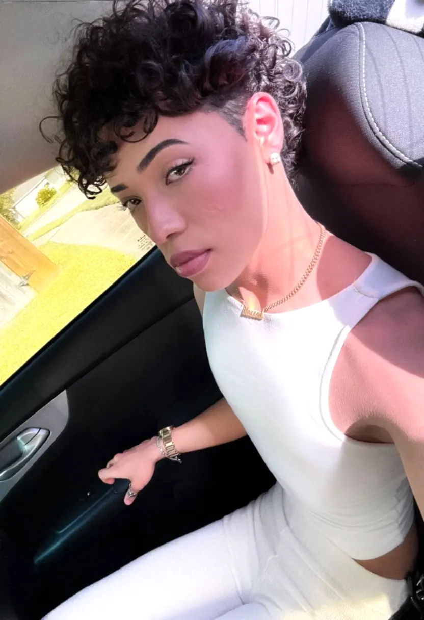 Person with short curly hair seated inside a car taking an angled close-up selfie.